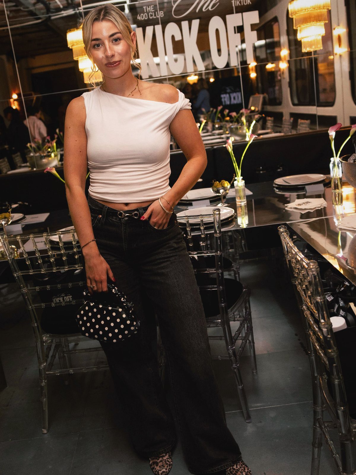 A woman wearing a white one-shoulder top and black wide-leg jeans posing in a restaurant.