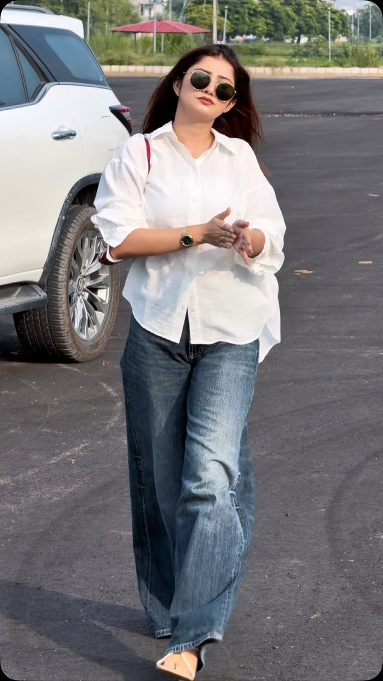 A woman in a white shirt and wide leg jeans posing outdoors next to a car.