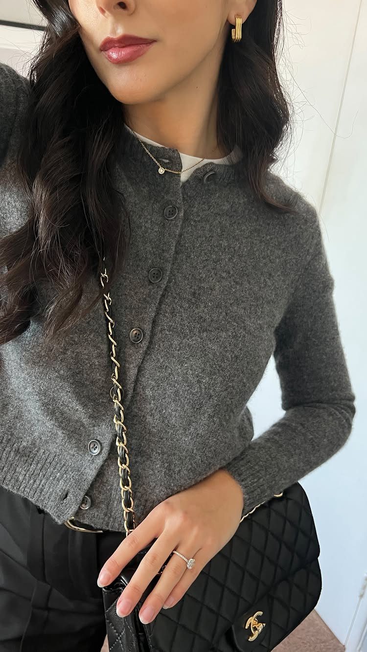 A woman wearing a grey knit button-up cardigan and black trousers with a quilted handbag.