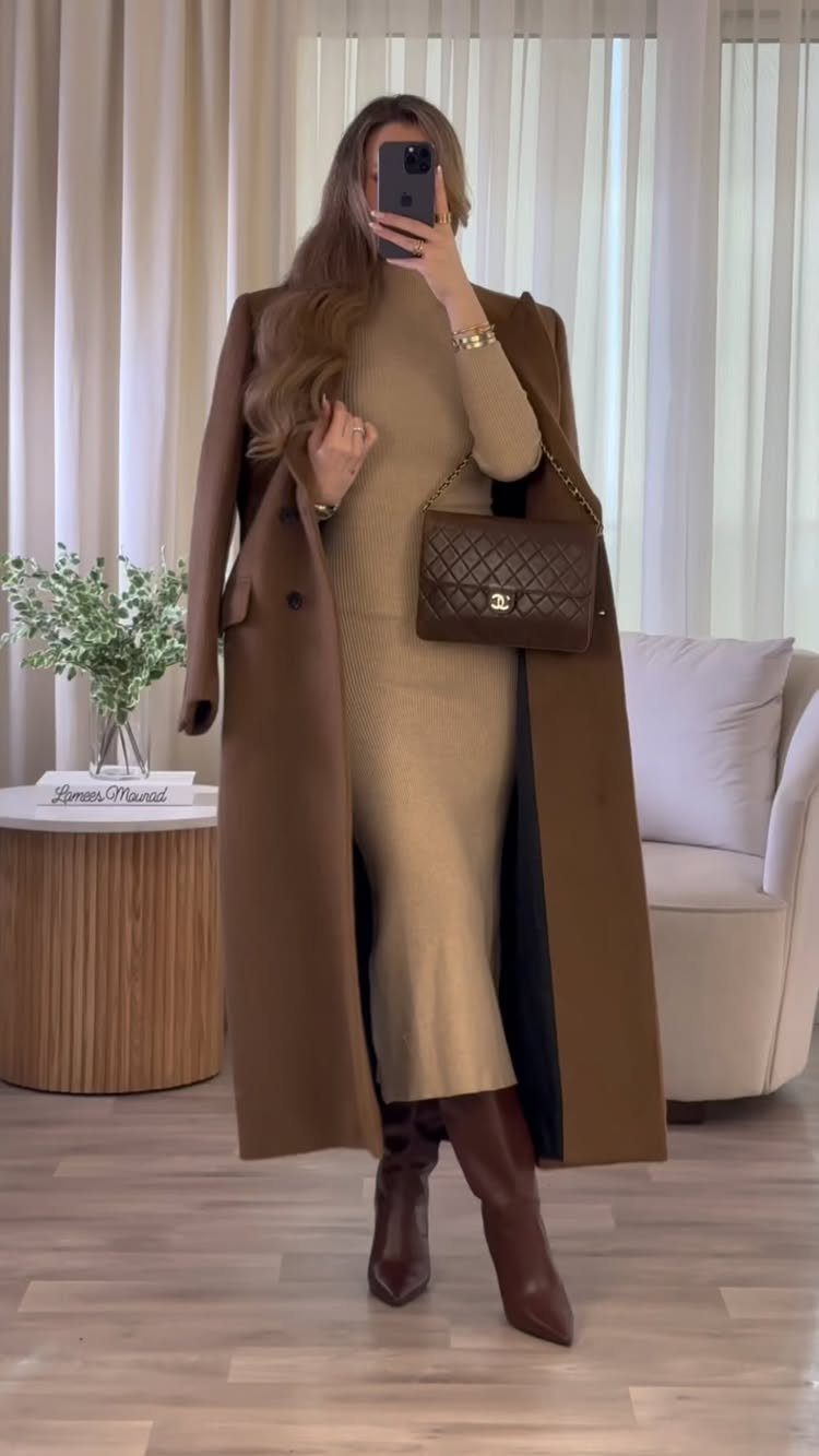 Woman in a tan knit dress, brown coat, and leather boots posing for a mirror selfie.