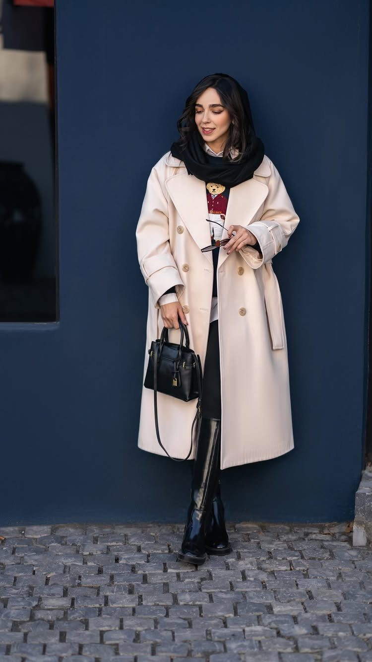 A woman in a cream overcoat, black scarf, and black boots standing against a blue wall.