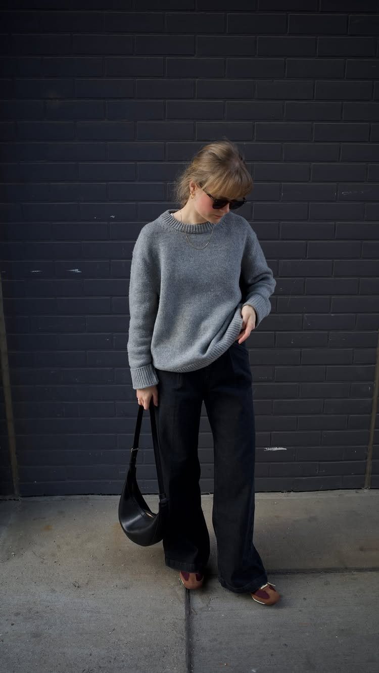 Woman wearing a grey crewneck pullover sweater and wide-leg black trousers standing against a dark wall.