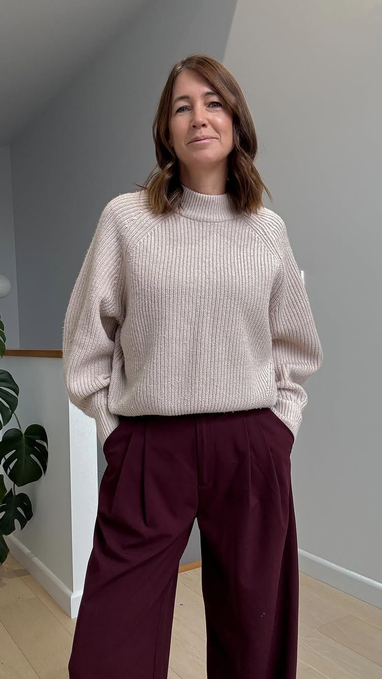 A woman wearing a cream knit sweater and burgundy wide-leg tailored trousers standing indoors.