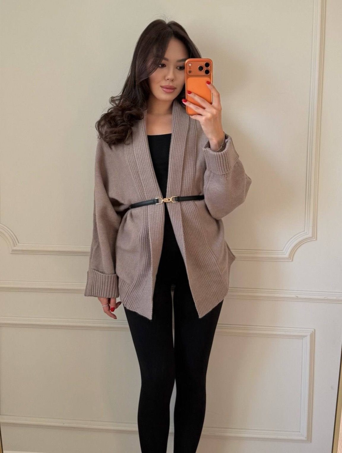 A woman posing in a beige ribbed cardigan and black leggings against a white wall.
