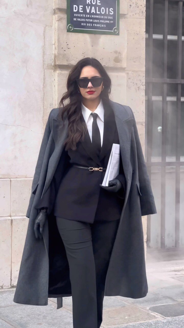 Woman in a black suit, white shirt, and black tie walking outdoors with a grey coat.
