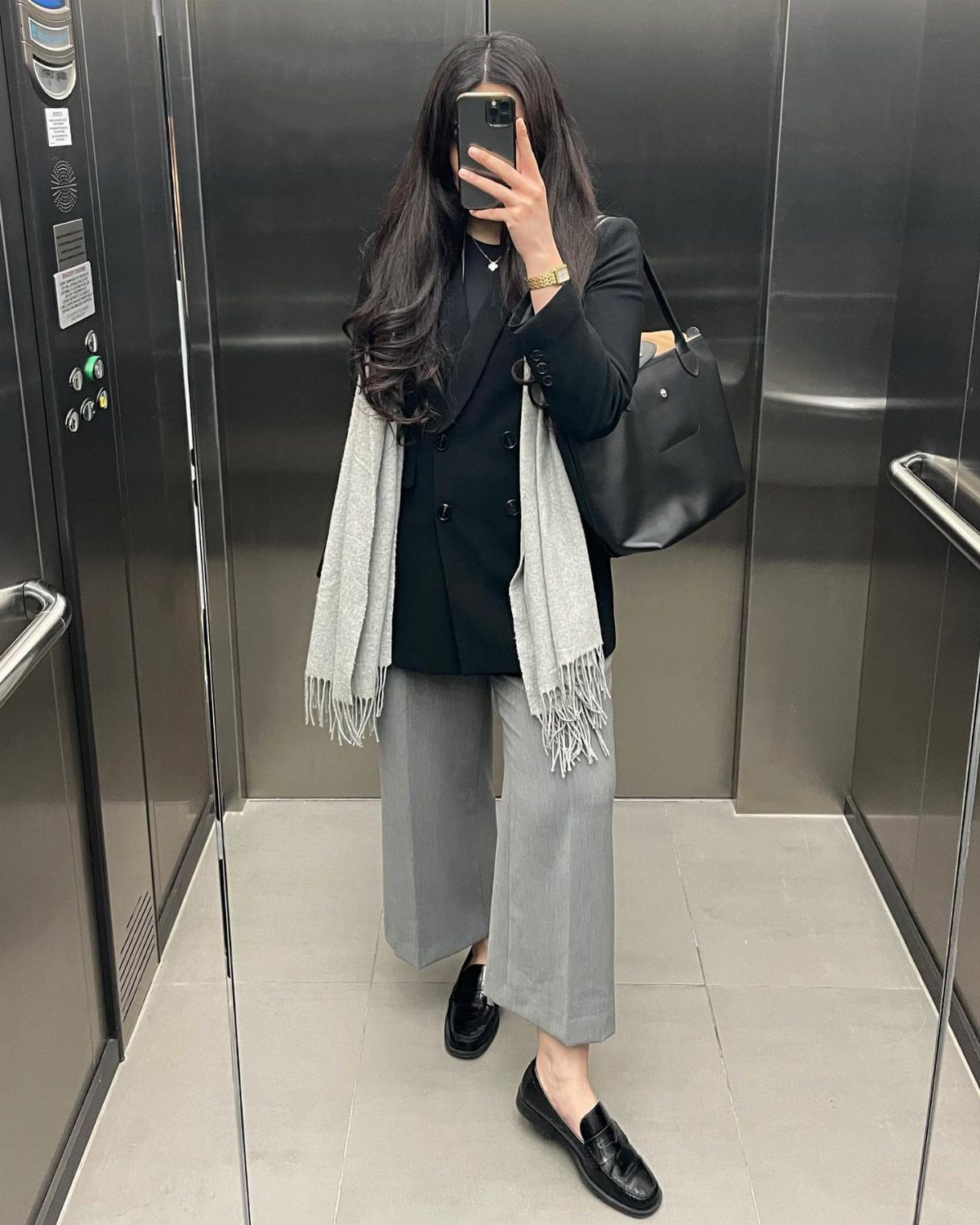 A person wearing a black blazer, grey trousers, and black loafers standing in an elevator.