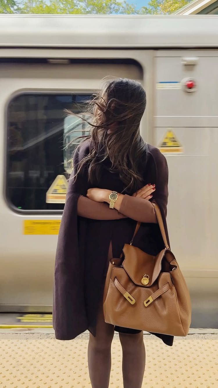 Woman in a dark purple cape holding a large tan leather handbag in front of a train.