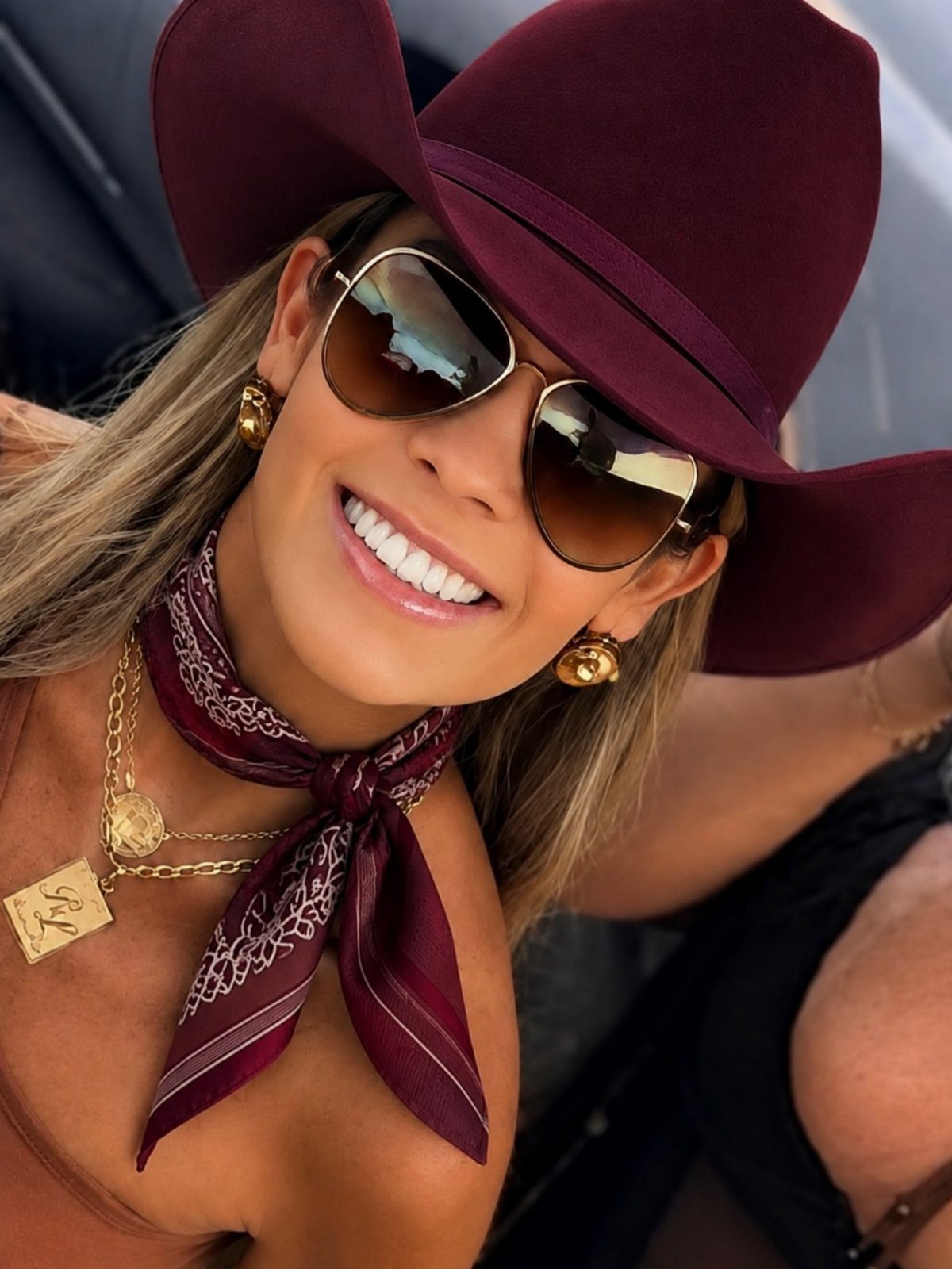 Smiling woman wearing large aviator sunglasses, a burgundy wide-brimmed hat, and a patterned neck scarf.