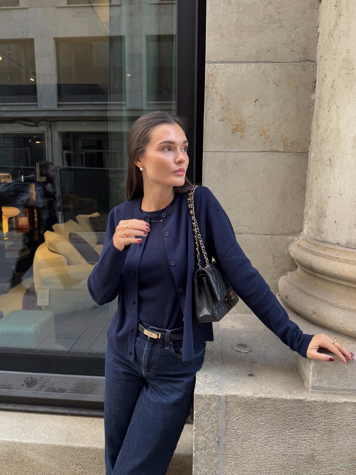 A woman wearing a navy blue cardigan and dark jeans posing by a stone pillar.
