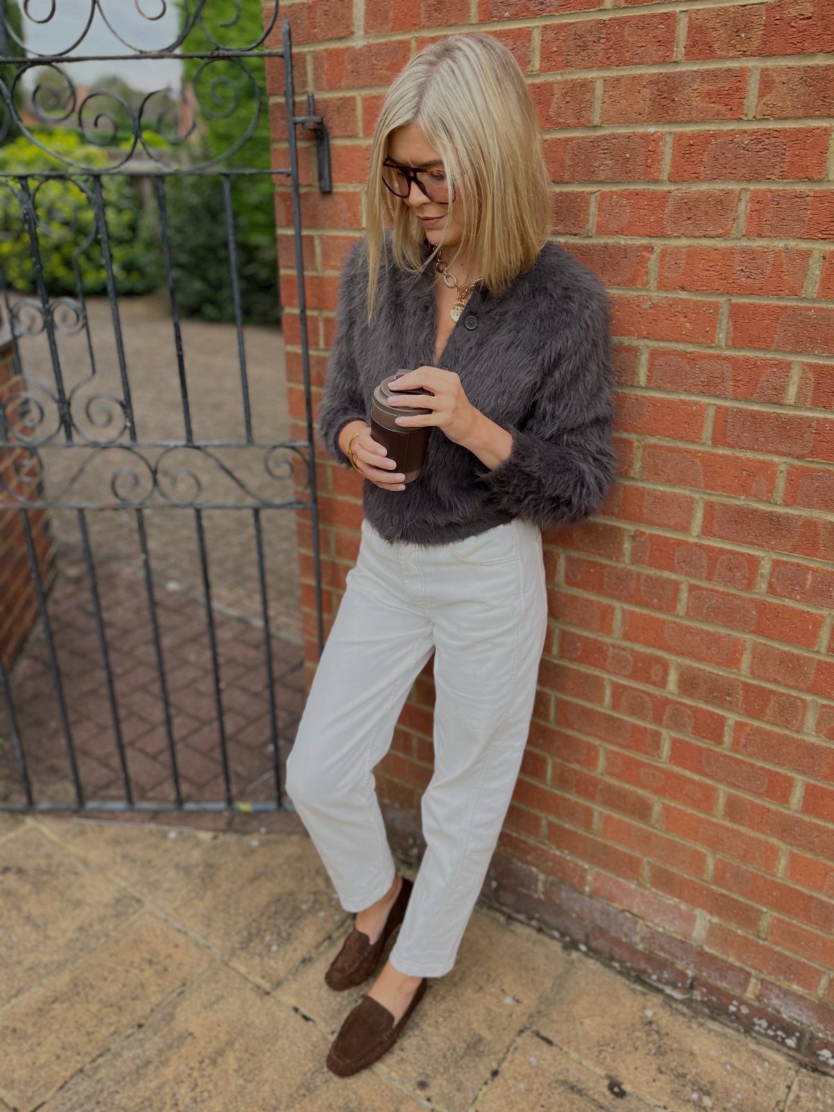 Blonde woman in white trousers and brown loafers leaning against a red brick wall.