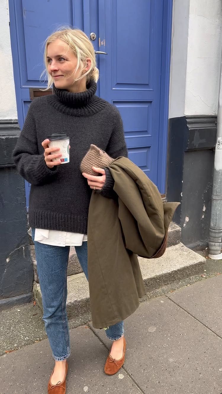 Blonde woman wearing a dark turtleneck, blue jeans, and brown ballet flats standing outdoors.