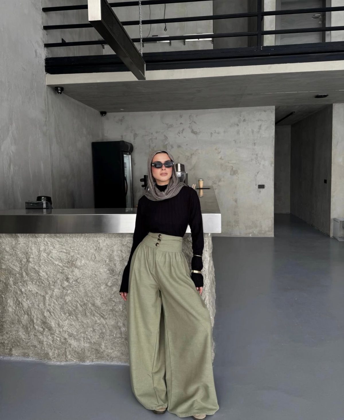 Woman posing in black top and wide-leg olive green trousers in a minimalist room.