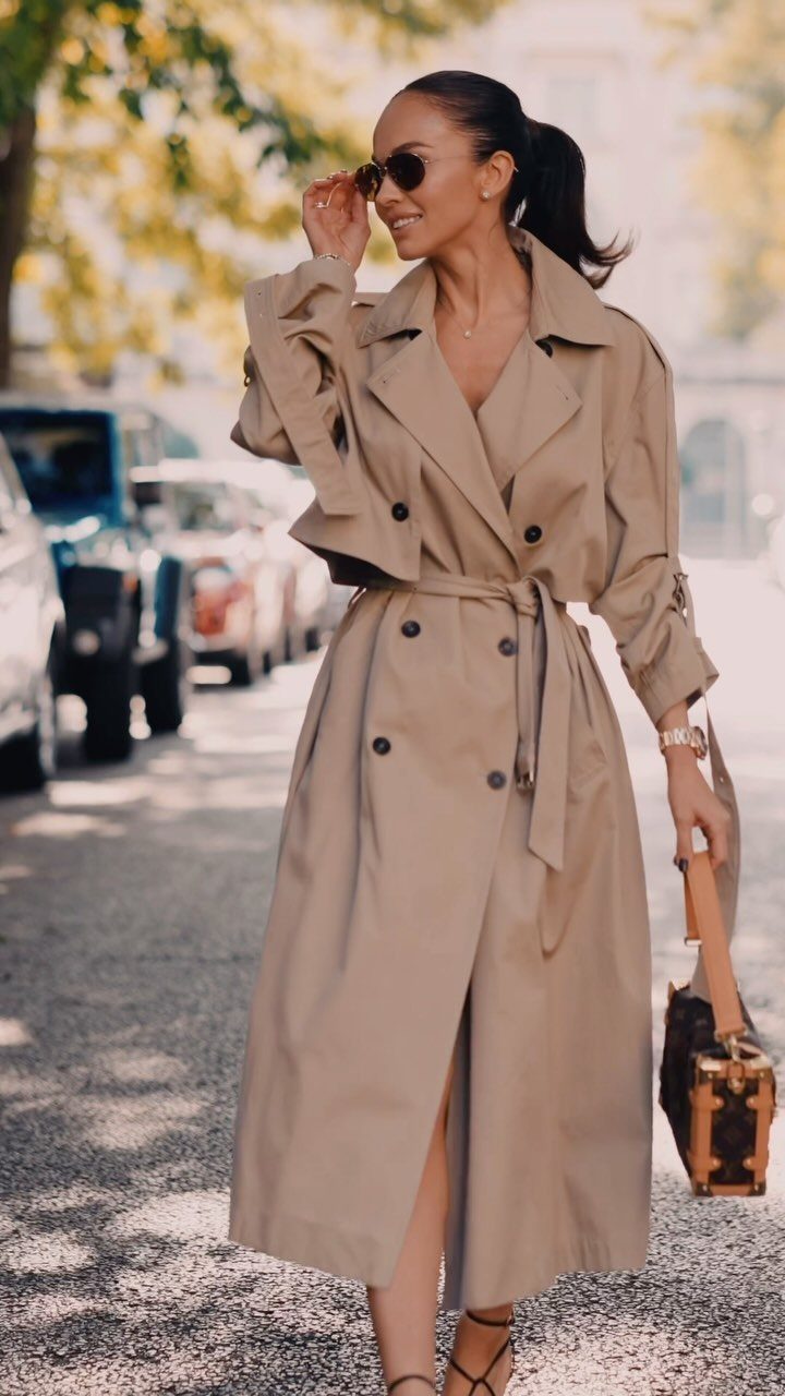 A woman walking down a city street wearing a long tan belted trench coat and sunglasses.