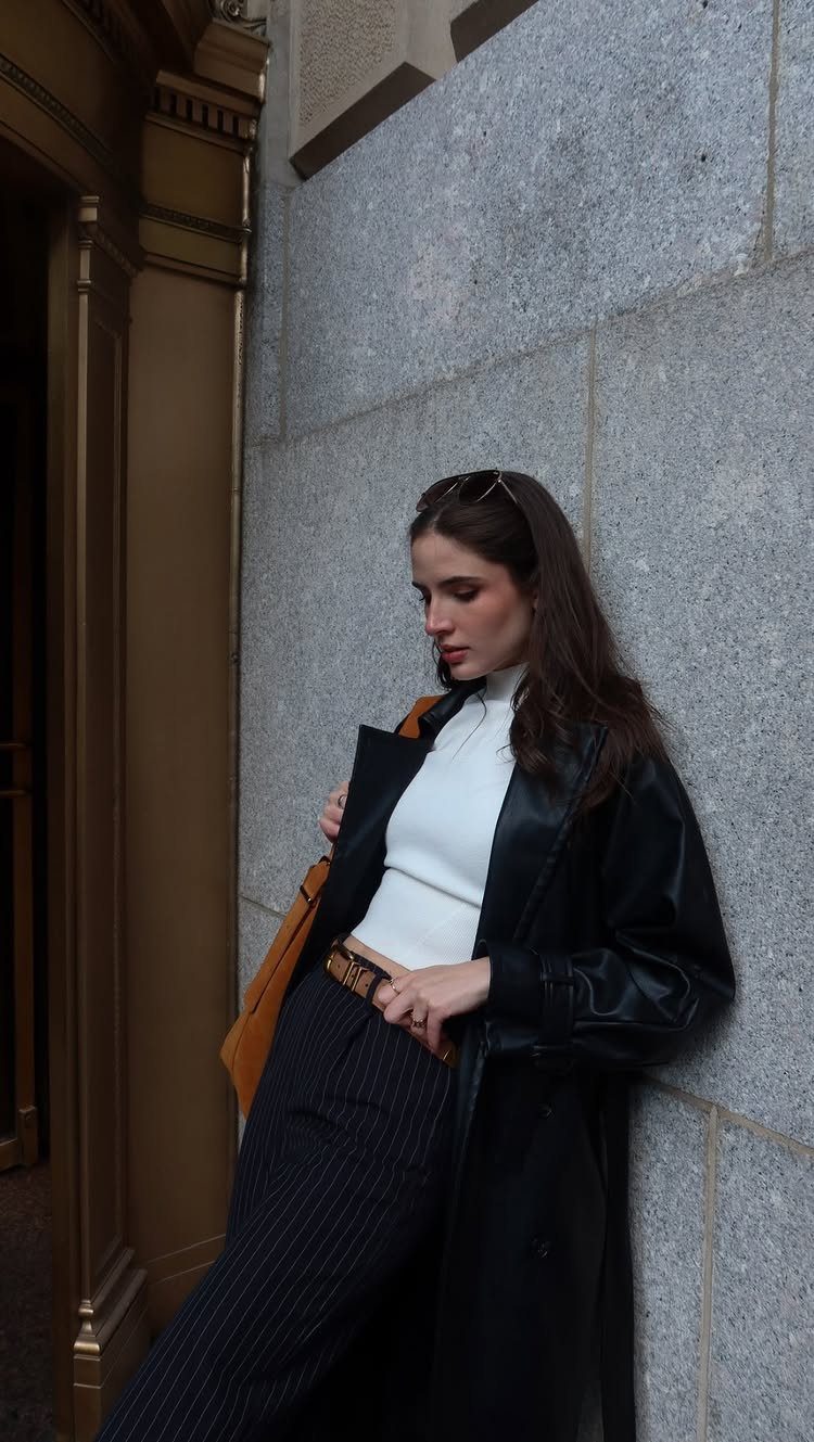 Woman in a black leather coat, white turtleneck, and pinstripe trousers leaning against a stone wall.