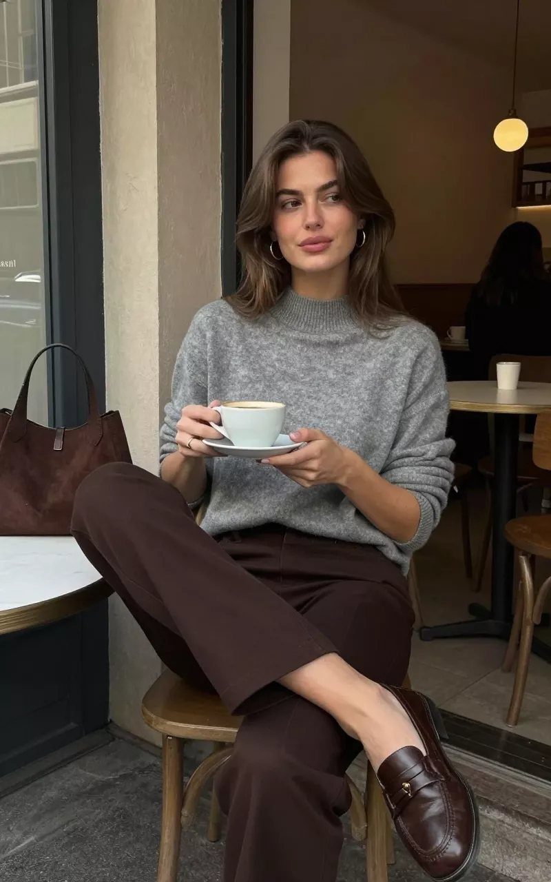 A woman in a grey sweater and brown trousers wearing leather loafers while sitting at a cafe.