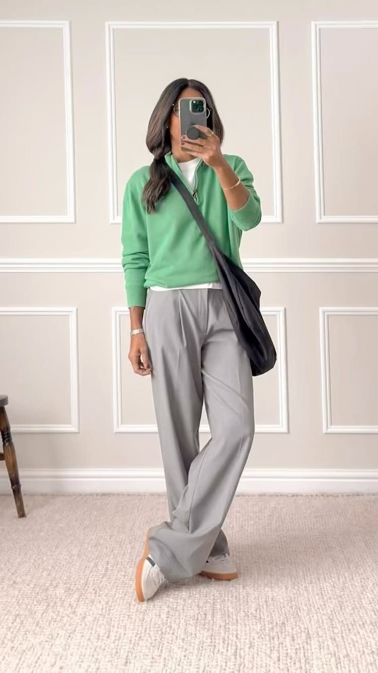 A woman posing in a green sweater and grey wide-leg trousers for a mirror selfie.