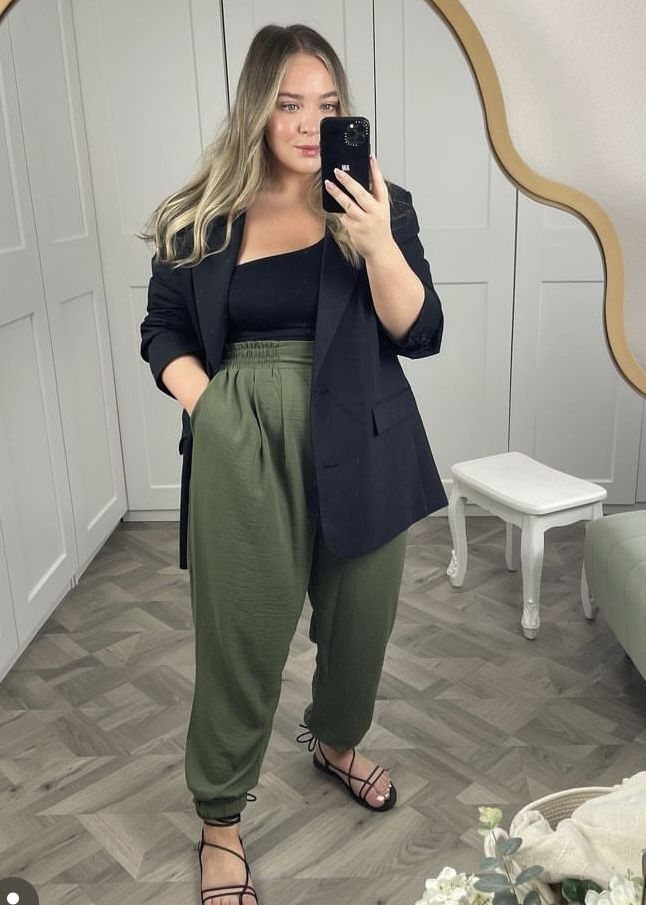 A woman posing in a black blazer, black tank top, and olive green joggers while taking a mirror selfie.