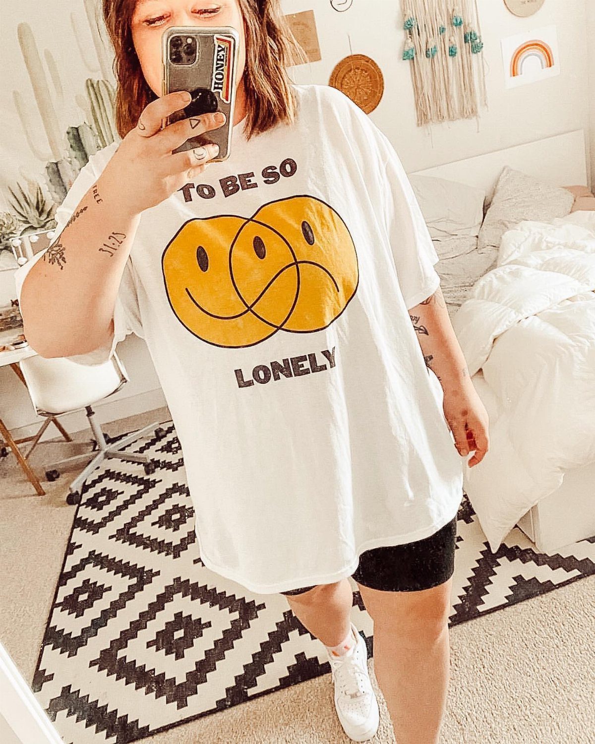 A person wearing an oversized white graphic t-shirt with yellow smiley faces and black shorts.