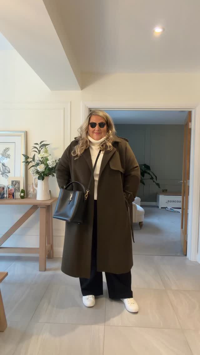 A woman wearing an olive green trench coat, white turtleneck, and black wide-leg trousers.